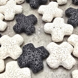 May include: Close-up of several cross-shaped decorative stones. The stones are in shades of white and dark grey, with a porous texture. The crosses are scattered across a light grey surface.