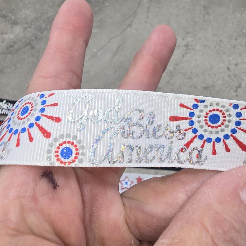 May include: White ribbon with a patriotic design featuring red, white, and blue accents. The ribbon has a repeating pattern of stylized starbursts and the text "God Bless America" in a shimmering silver font.