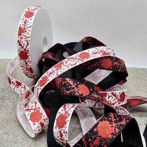 May include: A roll of white ribbon with a red splatter design sits next to a pile of black and white ribbons with a similar pattern. The red splatters have a glittery texture. The ribbons are likely for crafting or gift wrapping.