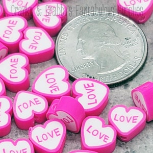 Könnte beinhalten: Rosa und weiße herzförmige Perlen mit dem Wort "LOVE" darauf. Die Perlen sind etwa so groß wie eine US-amerikanische 25-Cent-Münze.
