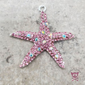 Op de afbeelding: Een zilveren zeester-bedel met roze iriserende strass steentjes. De bedel heeft een lus aan de bovenkant om aan een ketting of armband te bevestigen.