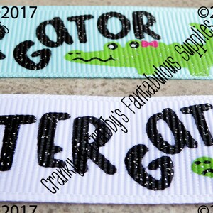May include: Two white and light blue ribbon trims with black glitter text that reads "LATER GATOR" and a green alligator with a pink bow. The text is on a white background and the alligator is on a light blue background.