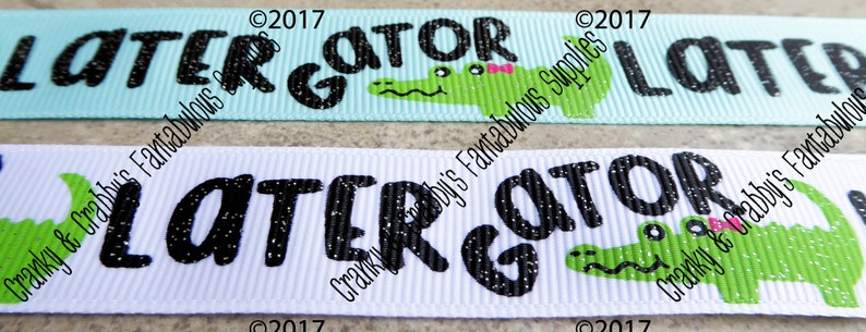 May include: Two white and light blue ribbon trims with black glitter text that reads "LATER GATOR" and a green alligator with a pink bow. The text is on a white background and the alligator is on a light blue background.