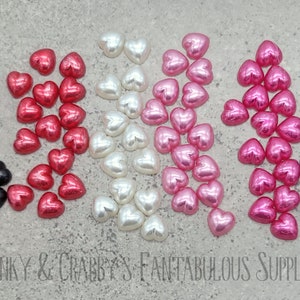 Op de afbeelding: Assortiment van hartvormige kralen in verschillende kleuren, waaronder zwart, rood, wit, roze en paars. De kralen zijn in rijen op een grijs oppervlak gerangschikt.