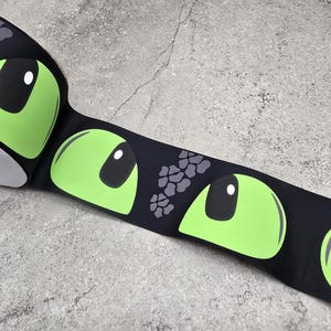 Può includere: Un nastro in grosgrain nero caratterizzato da un motivo grafico ripetuto di occhi alieni verde lime brillante con pupille nere e riflessi bianchi, intervallati da piccoli gruppi di macchie testurizzate grigio scuro lungo il tessuto.