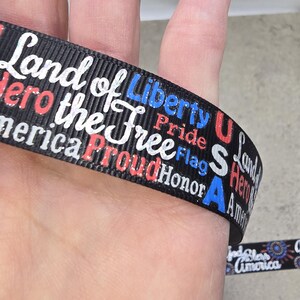May include: Black ribbon with patriotic text in white, red, and blue. The ribbon features phrases like "Land of Liberty," "Proud," and "America." The ribbon is wrapped around a finger, showcasing the design.