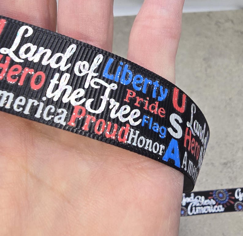 May include: Black ribbon with patriotic text in white, red, and blue. The ribbon features phrases like "Land of Liberty," "Proud," and "America." The ribbon is wrapped around a finger, showcasing the design.
