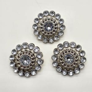 Puede incluir: Tres adornos plateados en forma de flor con brillantes pedrería transparente y brillante. Cada uno tiene una piedra central grande rodeada de piedras más pequeñas, creando un efecto deslumbrante. Ideal para añadir brillo a la ropa o accesorios.
