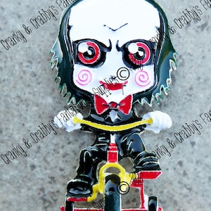 May include: A metal charm featuring a cartoon character with a white face, red eyes, and a red bow tie, riding a red and yellow tricycle. The character has black hair and is wearing a black suit. The charm has a loop at the top for hanging.