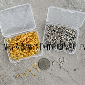 以下が含まれることがあります： 金と銀のアイピンが詰まった透明なプラスチック容器が2つ。容器はグレーのテクスチャのある表面の上に置かれています。容器の周りにアイピンがいくつか散らばっています。