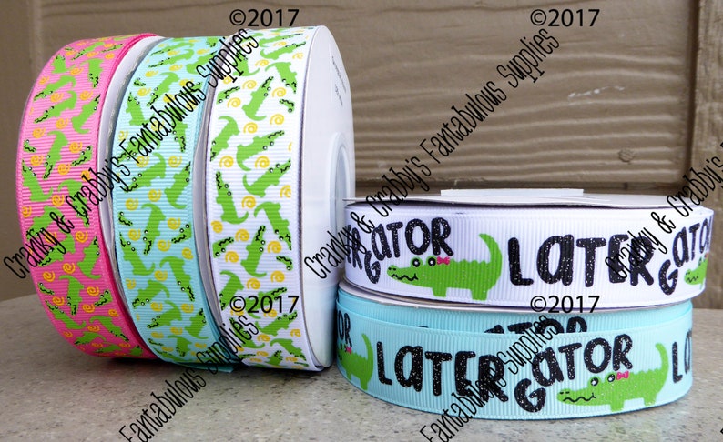 May include: Four rolls of white satin ribbon with alligator designs. The ribbon is printed with green alligators and yellow circles. One roll has the text "Gator" with a green alligator and a pink bow. Another roll has the text "Later Gator" with a green alligator and a pink bow. The ribbon is 2.5 inches wide.