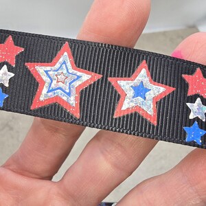 May include: Black ribbon featuring a repeating pattern of red, white, and blue stars. The stars have a glittery finish and are arranged in a patriotic design. The ribbon is held in a hand.