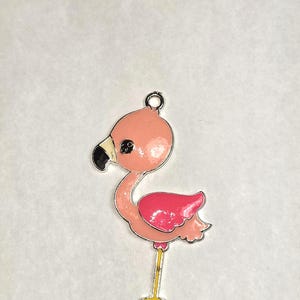 May include: A pink flamingo charm with a silver-colored outline. The flamingo has a black beak, a pink body, and a darker pink wing. The charm has a small loop at the top for attaching to a necklace or bracelet.