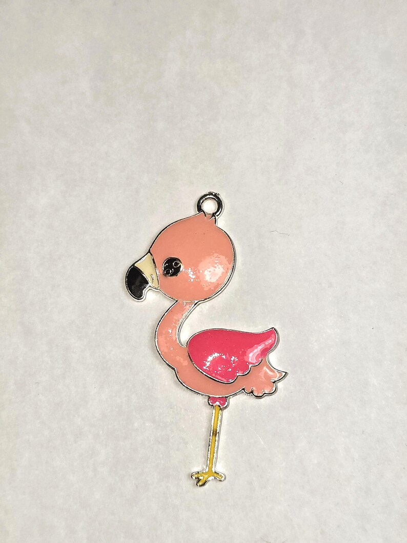 May include: A pink flamingo charm with a silver-colored outline. The flamingo has a black beak, a pink body, and a darker pink wing. The charm has a small loop at the top for attaching to a necklace or bracelet.