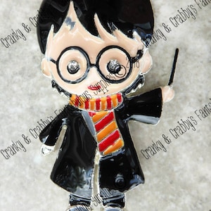 May include: A black and silver charm of Harry Potter, wearing a black robe with a red and orange striped scarf, holding a wand in his right hand. He is wearing round glasses with rhinestones for eyes.
