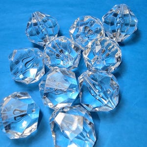 May include: A collection of clear, faceted crystal beads. Each bead has multiple geometric faces that reflect light, creating a sparkling effect. The beads are scattered on a solid blue background.