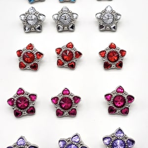 May include: A collection of twelve star-shaped decorative buttons. Each button features a central gemstone surrounded by smaller heart-shaped accents. The buttons come in various colors, including blue, clear, red, pink, and purple.
