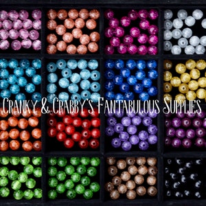 May include: A collection of 12 different colors of round, glittery beads in a black tray. The beads are arranged in a grid pattern. The text "CRANKY & CRABBY'S FANTABULOUS SUPPLIES" is printed on the tray.