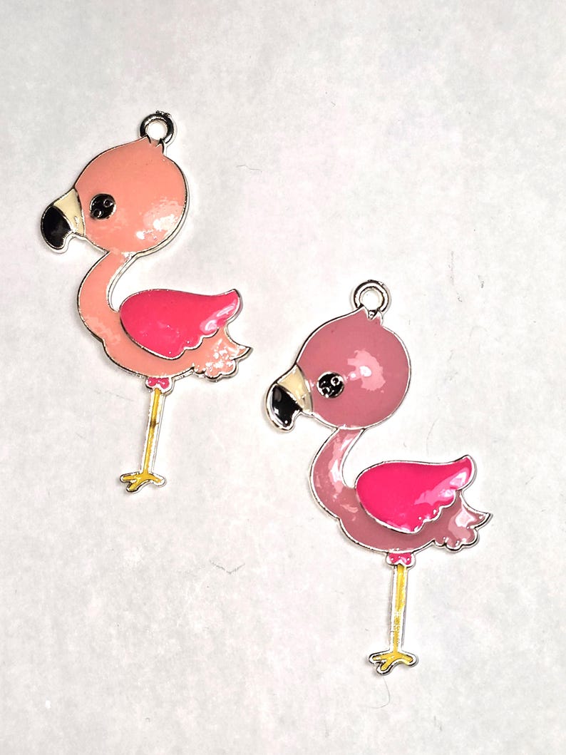 May include: Two flamingo charms with pink and fuchsia enamel. The charms have black beaks, yellow legs, and silver-colored metal trim. Each charm has a loop at the top for attaching to jewelry or other crafts.