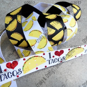 May include: Two rolls of white ribbon with a black and white taco print and a red heart with the text "I love tacos". A third roll of black ribbon with a white taco print is on top of the other two rolls.