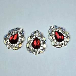 May include: Three ornate, teardrop-shaped brooches. Each brooch features a large, faceted, red gemstone at the center, surrounded by smaller clear and iridescent crystals. The setting is a gold-toned metal.