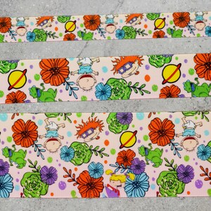 May include: Pink ribbon with a repeating pattern of cartoon characters, flowers, and planets. The characters are from the Nickelodeon show Rugrats.
