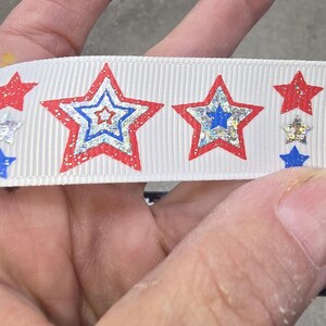May include: White ribbon featuring a repeating pattern of red, blue, and silver stars. The stars have a glittery, metallic appearance, with a red outline and a blue or silver center. The ribbon is likely for crafting or decorative purposes.