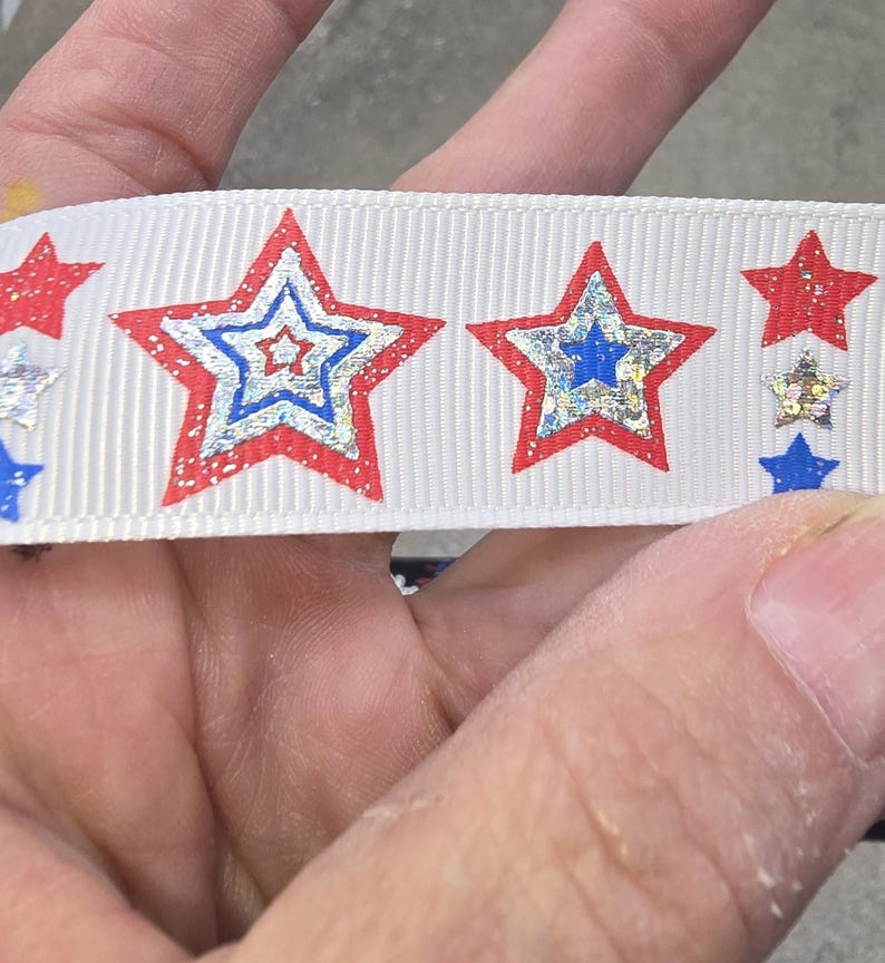 May include: White ribbon featuring a repeating pattern of red, blue, and silver stars. The stars have a glittery, metallic appearance, with a red outline and a blue or silver center. The ribbon is likely for crafting or decorative purposes.