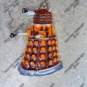 May include: A silver and orange Dalek ornament with a detailed design. The Dalek is a fictional robot from the British science fiction television series Doctor Who.