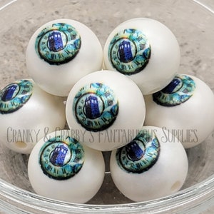 May include: A close-up of white round beads with a blue and green image of a clock and a blue police box. The beads are in a clear glass bowl. "CRANKY & GRABBY'S FANTASTIC SUPPLIES" is printed on the bottom of the bowl.