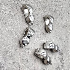May include: A collection of five shiny, silver-colored metal clasps. Each clasp has a curved, organic shape with a small loop at one end and a hinged closure at the other. The clasps are scattered on a textured, gray surface.