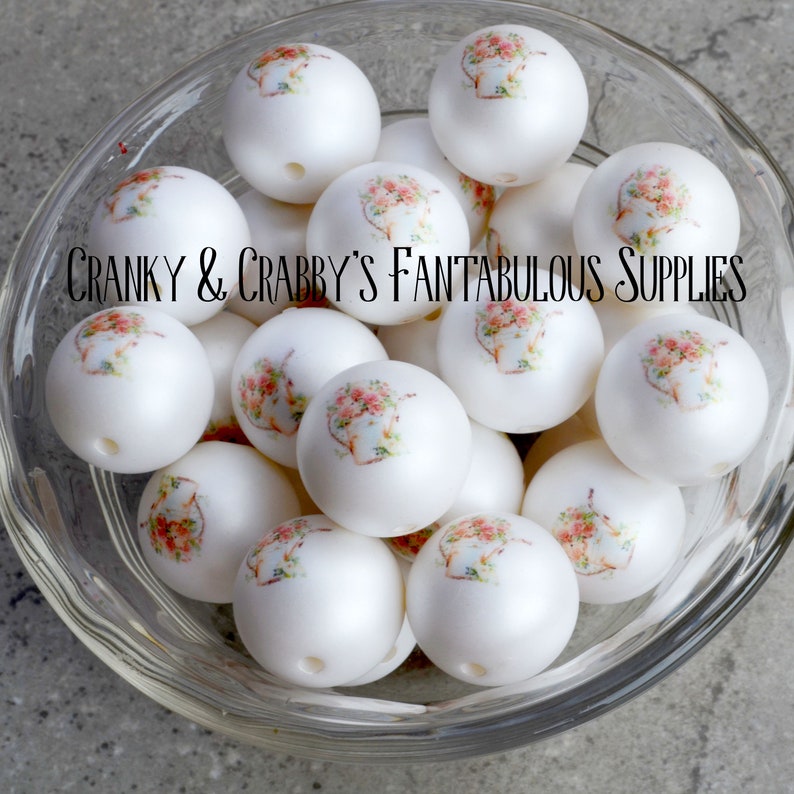 Pode incluir: Uma tigela de contas redondas decorativas brancas com um design floral. As contas t&ecirc;m um padr&atilde;o floral de inspira&ccedil;&atilde;o vintage com acentos rosa e verdes. O texto "Cranky & Crabby's Fantabulous Supplies" &eacute; vis&iacute;vel na imagem.