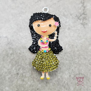 Op de afbeelding: Een bedel met een cartoon hula-meisje met zwart haar, een roze bloem en een groene en gele lei. De bedel is gemaakt van metaal en heeft een glanzende afwerking.