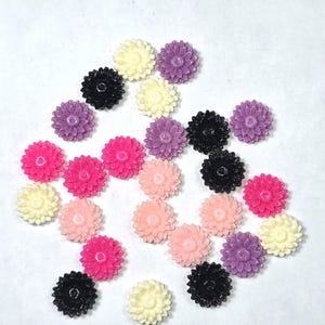 May include: A selection of small, daisy-shaped embellishments in various colours, including black, pink, purple, peach, and cream. These decorative pieces are scattered across a white surface, showcasing their detailed floral design.