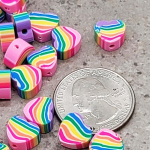 Könnte beinhalten: Nahaufnahme eines Stapels regenbogenfarbener herzförmiger Perlen. Die Perlen sind aus Polymer Clay gefertigt und haben eine rosa Basis mit Regenbogenstreifen. Die Perlen sind etwa so groß wie eine US-amerikanische 25-Cent-Münze.