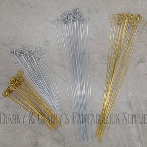 Peut inclure: Vue aérienne de trois paquets de brochettes décoratives. Deux sont argentées et une dorée. Chaque brochette est surmontée d'un cœur. Le texte "Cranky & Graby's Fantabulous Supplies" est visible en bas à gauche.