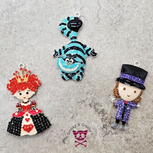 Puede incluir: Tres colgantes de charms con personajes de Alicia en el País de las Maravillas. El primer charm es una Reina de Corazones roja y negra con una corona. El segundo charm es un Gato de Cheshire azul y negro. El tercer charm es un Sombrerero Loco morado y negro con un sombrero de copa.