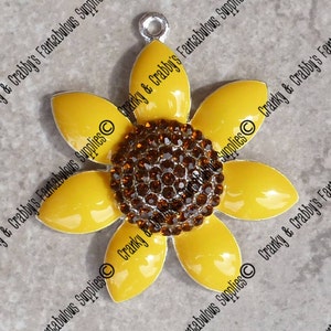 May include: A yellow sunflower charm with a brown rhinestone center. The charm has a silver loop for attaching to a necklace or bracelet.