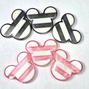 Può includere: Cinque abbellimenti in resina a forma di Topolino. Tre sono a righe bianche e nere e due a righe rosa e bianche. Gli abbellimenti hanno una finitura lucida e sono disposti su una superficie bianca.