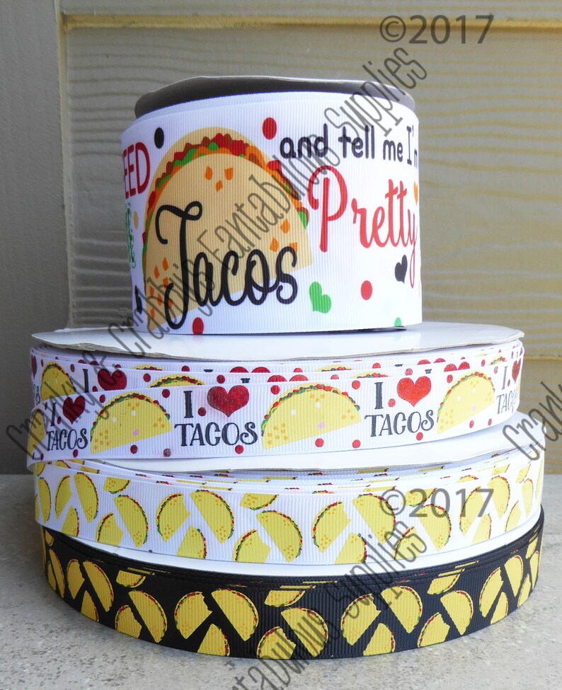 May include: A stack of four white and black ribbon rolls with a taco theme. The top ribbon has the text "and tell me I'm pretty tacos" printed on it. The second ribbon has the text "I love tacos" printed on it. The third ribbon has a pattern of yellow tacos on a white background. The bottom ribbon has a pattern of yellow tacos on a black background.