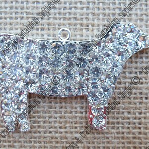 May include: A silver cow charm covered in clear rhinestones. The charm has a loop at the top for attaching to a necklace or bracelet.