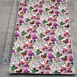 May include: A white fabric sheet with a repeating pattern of Mickey Mouse, Minnie Mouse, ghosts, pumpkins, and stars. The pattern is in shades of orange, green, purple, and pink.