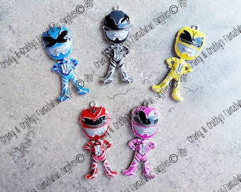 Könnte beinhalten: Fünf Metall-Anhänger mit Power Rangers-Charakteren. Die Anhänger sind in den Farben Schwarz, Blau, Gelb, Rot und Pink. Jeder Anhänger zeigt einen anderen Charakter mit einem gestreiften Muster.