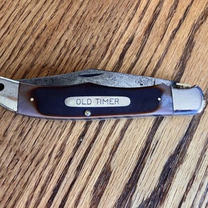 May include: A closed folding pocket knife with a dark brown handle and a silver-colored metal bolster. The handle features a central silver-colored inlay with the text OLD TIMER engraved in capital letters. The metal blade is partially visible.