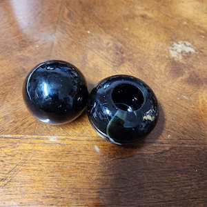 May include: Two black glass spheres sit on a wooden surface. One sphere is solid, while the other has a hollowed-out center. The glass has a glossy finish, reflecting light. The spheres are likely decorative objects.