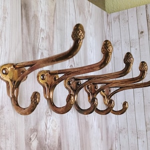 May include: A set of four brass-toned metal wall hooks, each featuring a double-pronged design with a curved upper arm and a smaller lower hook. The ends of each prong are finished with a textured, acorn-shaped decorative finial.
