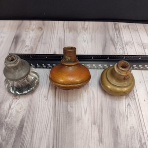 May include: Three door knobs are arranged in a row next to a black ruler. The left knob is clear glass with a fluted design, the center knob is a smooth brown metal, and the right knob is a smooth brass metal. Each knob has a threaded neck.