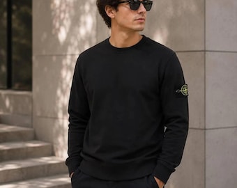 May include: A black long-sleeve crewneck sweatshirt featuring a circular yellow, green, and black compass-style patch logo on the left sleeve. The garment has a ribbed neckline, cuffs, and hem, providing a clean and structured appearance.