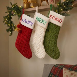 Peut inclure: Trois chaussettes de Noël en tricot torsadé suspendues. La chaussette rouge porte le nom LAURY en violet, la blanche porte MERY en vert, et la verte porte le mot STUDIO en rouge sur des revers blancs brodés de manière distincte.