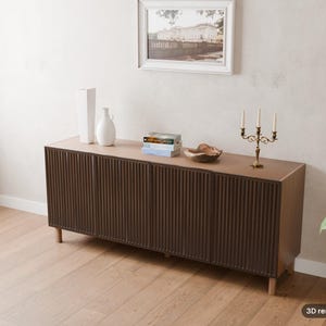 May include: A long, dark brown wooden sideboard featuring four cabinet doors with a vertical ribbed texture. The sideboard stands on light-coloured wooden legs and is topped with two white vases, a stack of books, a wooden bowl, and a gold candelabra.